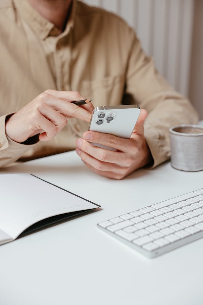 Person interacts with smartphone using a stylus at a clean, white office desk with keyboard and notepad.